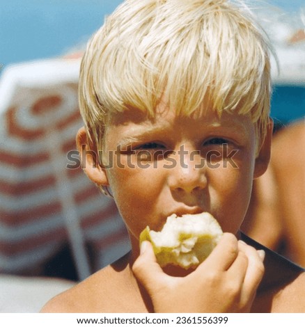Vintage roll film retro 1979 image, close-up portrait of a young boy with blond hair and beach umbrella background eating a green apple. Royalty-Free Stock Photo #2361556399