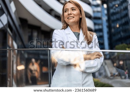 Smiling Caucasian female healthcare worker with arms crossed. A young, confident doctor outside a large hospital.