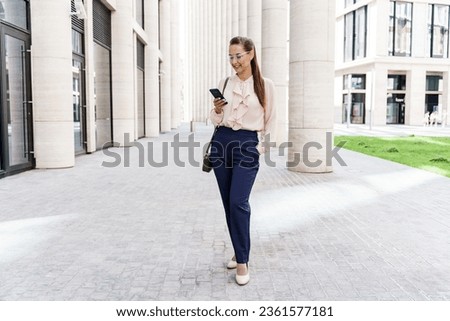 A modern woman in office clothes, a bank employee. A business woman uses a phone, writes a message to a client. The financier is standing near an office building.