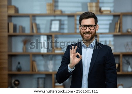 OK sign, businessman shows approval sign to camera, smiling man in business suit inside office at workplace, experienced financier investor in office.