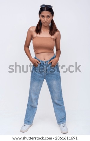 A fierce Filipino woman in her late teens or early 20s. Wearing a brown top and loose fitting jeans. Isolated on a white backdrop. Royalty-Free Stock Photo #2361611691