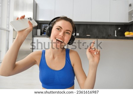 Close up portrait of happy fitness girl, listens music in wireless headphones, holds smartphone, dances and workout from home.