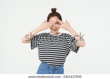 Happy young woman, bday girl peeking through fingers, holds hands on eyes, waiting for surprise, stands over white background.