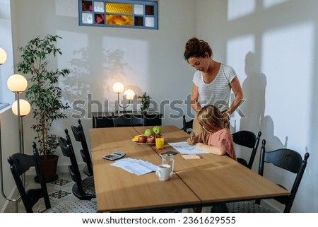 Caucasian mother and daughter in a modern room, mother is watching her daughter drawing while she is feeling proud