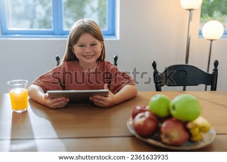 Little blonde girl sitting at the dining table and smiling for the camera while using her tablet