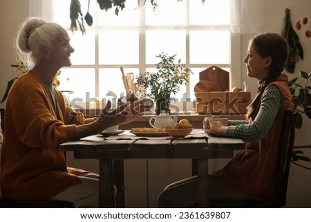 Happy grandmother and granddaughter sitting by wooden table against window in the kitchen, having tea with homemade food and chatting Royalty-Free Stock Photo #2361639807