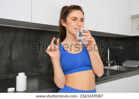 Portrait of beautiful, brunette woman, wearing fitness activewear, drinking water with dietary supplement, taking vitamin D buds for healthy, strong body, standing in kitchen.