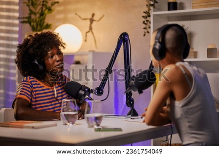 Two co-hosts are sitting in a home recording studio and discussing topics while wearing headphones and speaking at the microphone. Podcasters live stream on air for social media and make a podcast.