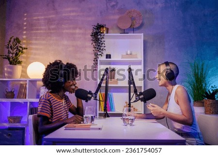 Two happy multiracial young women are sitting in a podcast studio and live-streaming their discussion. Gen z girls are making a podcast. Two women wearing headphones and talking at the microphone. Royalty-Free Stock Photo #2361754057