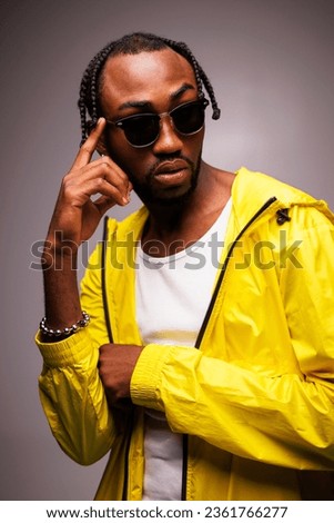 Portrait of stylish african american man with braids wearing sunglasses, tank top and windbreaker jacket isolated on grey