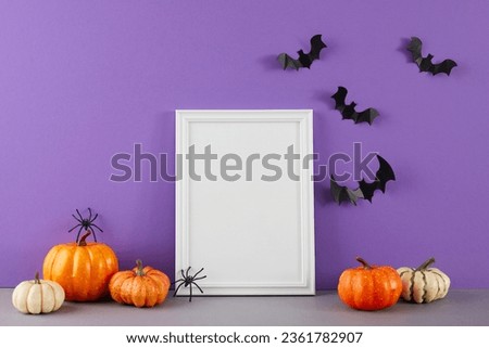 Emerging fully in the supernatural atmosphere that surrounds Halloween. Side view of colorful pumpkins on the table, spooky bats and spiders on purple background with empty frame for note or ads