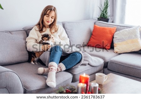 Woman in plaid with tea cup watching movie, TV, petting cat on sofa at home with christmas decoration atmosphere. Lady wear jumper and warm socks. Cozy and comfortable winter concept. Selective focus.
