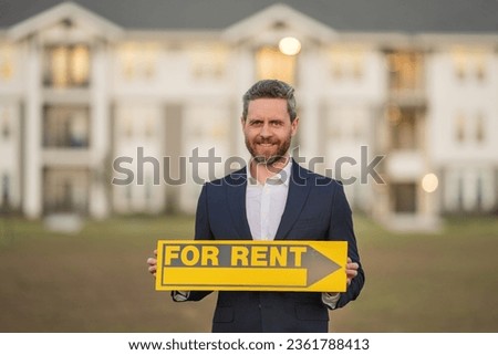 Successful real estate agent in a suit holding for rent sign near new apartment. Real estate agent with rental contract, renting home. Realtor or real estate agent shows board for rent. Rental house.