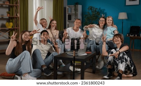 Full-size shot of a group of teens, young people, friends sitting around the table, using a smartphone on a tripod to take a picture, photo together, showing peace gesture.