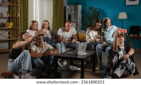 Full-size shot of a group of teens, young people, friends sitting on a couch and floor around the table, using their smartphones, taking selfies, pictures.
