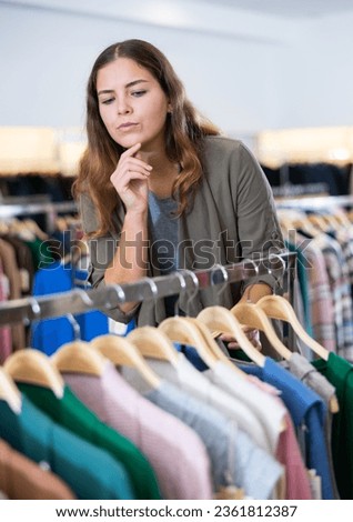 Serious young woman choosing season outfit clothes among the racks in a shopping mall. High quality photo