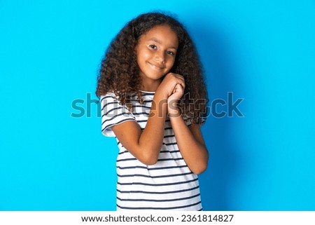Charming serious beautiful kid girl with afro curly hairstyle wearing striped T-shirt  keeps hands near face smiles tenderly at camera