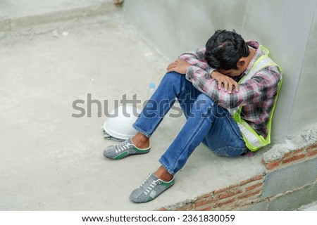 Discouraged construction worker sitting with back against cement wall at construction site. Stress because work is not completed on time. man wearing green reflective vest wears helmet to cool down. Royalty-Free Stock Photo #2361830059
