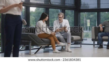 Asian woman sits on sofa in clinic lobby area after appointment with doctor. Doctor talks with patient about medical examination or test results. Waiting room in modern medical center. Healthcare. Royalty-Free Stock Photo #2361834381