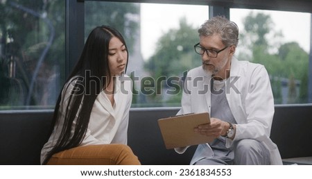 Asian woman sits on couch in clinic lobby area after appointment with doctor. Doctor talks with patient about medical examination or test results. Waiting room in medical center. Healthcare system.