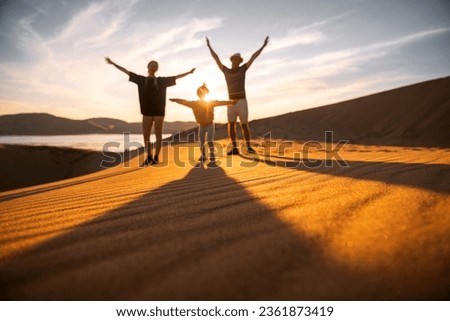 Silhouettes of happy family with little daughter are standing with open arms at sunset sea beach of sand dunes. Blurred silhouettes of unrecognizable family with raised arms