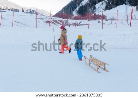 two children pulling the wooden sledge on snow slope in bright winter clothes