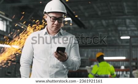 Multicultural industrial worker is walking at workshop. Male builder is holding smartphone. Asian engineer at background of work process. Man in white shirt and safety helmet at construction site.