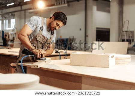 Young carpenter sanding wood piece in workshop in furniture factory Royalty-Free Stock Photo #2361923721