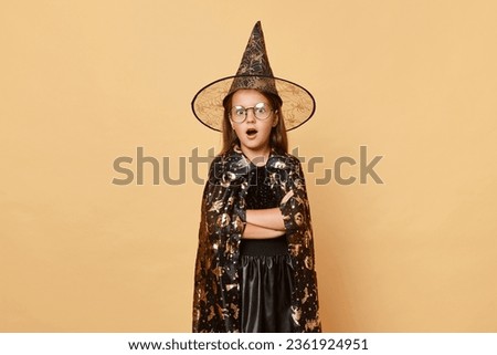 Shocked astonished surprised female kid girl wearing witch black hat costume isolated over beige background looking at camera with open mouth and big eyes halloween holiday.