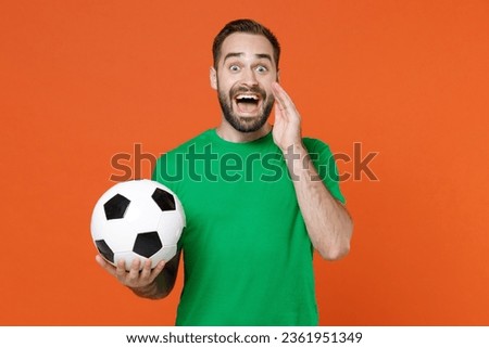 Excited man football fan in green t-shirt cheer up support favorite team with soccer ball screaming with hand gesture near mouth isolated on orange background. People sport leisure lifestyle concept