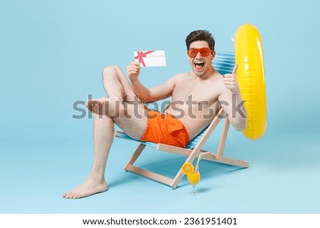 Excited young man guy in orange shorts glasses sit on deck chair isolated on blue background. People summer vacation rest lifestyle concept. Mock up copy space. Hold gift certificate showing thumb up