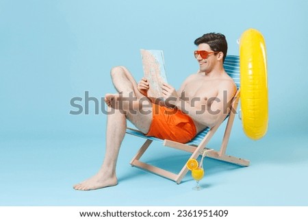 Smiling young man guy in orange shorts glasses sit on deck chair isolated on pastel blue background studio portrait. People summer vacation rest lifestyle concept. Mock up copy space. Hold city map