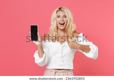 Excited young blonde woman 20s wearing white casual clothes pointing index finger on mobile phone with blank empty screen mock up copy space isolated on bright pink colour background, studio portrait