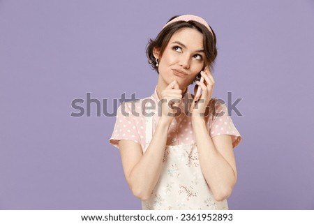 Young pensive housewife housekeeper chef cook baker woman in pink apron talking speaking by mobile cell phone prop p chin isolated on pastel violet background studio portrait. Cooking food concept. Royalty-Free Stock Photo #2361952893