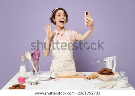 Young housewife housekeeper chef baker woman in pink apron work at table kitchenware do selfie shot by mobile cell phone waving hand isolated on pastel violet background Process cooking food concept.