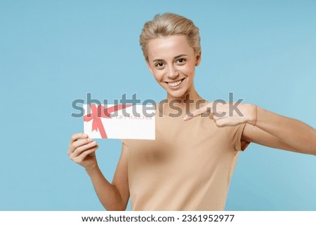Young smiling blonde woman short haircut dental braces in casual beige t-shirt hold point index finger on mockup of gift voucher certificate flyer isolated on blue color background studio portrait