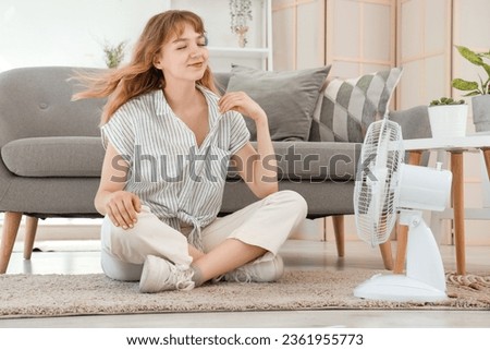 Happy beautiful young woman with electric fan sitting on floor in living room Royalty-Free Stock Photo #2361955773
