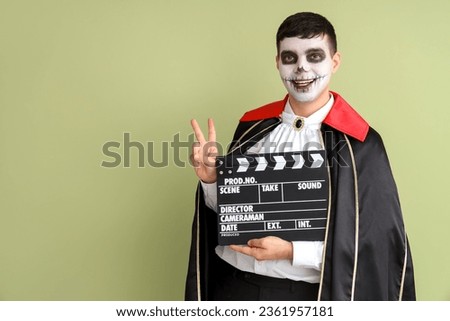 Young man dressed for Halloween as vampire with clapperboard showing victory gesture on green background