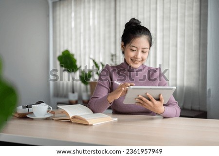 A happy and pretty Asian female college student using a digital tablet at a table in her living room. online study, online lecture, homework, internet connection Royalty-Free Stock Photo #2361957949