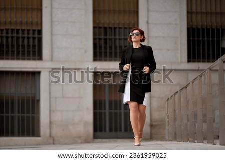 A sophisticated woman exudes elegance in a black and white dress with a jacket, accessorized with sunglasses while strolling through the city streets Royalty-Free Stock Photo #2361959025