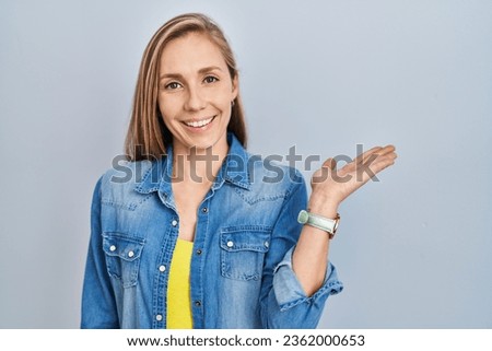 Young blonde woman standing over blue background smiling cheerful presenting and pointing with palm of hand looking at the camera. 