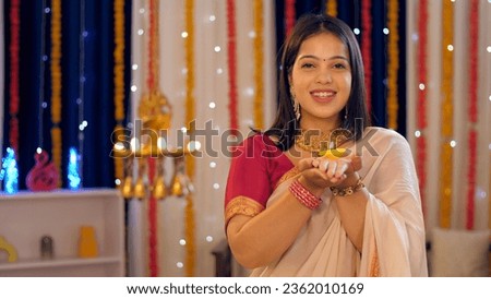 A beautiful woman holding a brightly burning clay diya to celebrate Diwali - greetings, posing for camera. A pretty lady in ethnic clothes posing for the camera against a festive background - India...