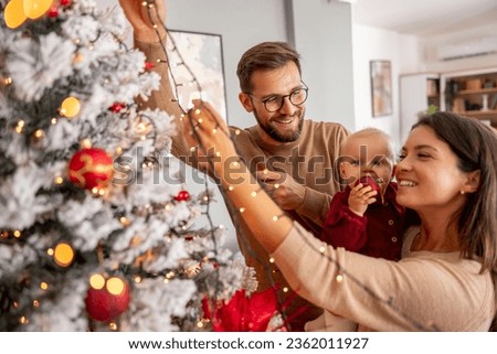 Young parents having fun decorating Christmas tree with their cute little baby girl, placing Christmas lights on it while decorating home for winter holiday season Royalty-Free Stock Photo #2362011927