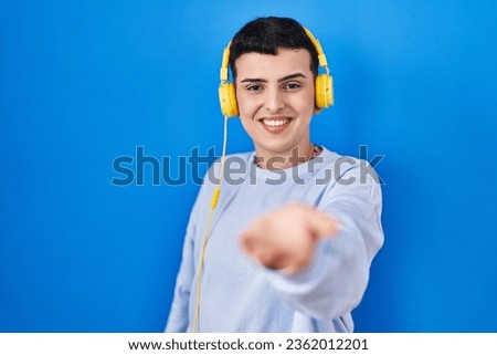 Non binary person listening to music using headphones smiling cheerful offering palm hand giving assistance and acceptance. 