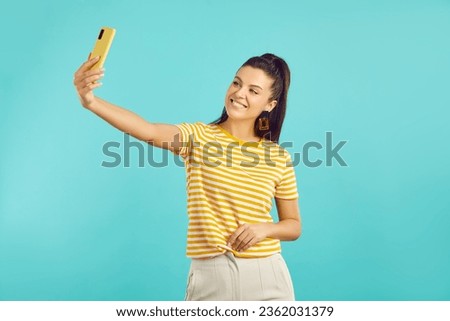 Happy young woman enjoys new mobile phone and takes a selfie on his camera on turquoise background. Beautiful cute caucasian woman in casual clothes looks at yellow phone in her hand. Banner.