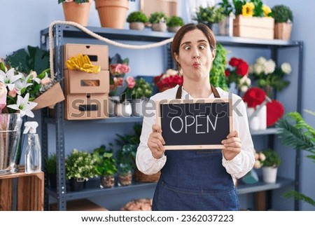 Brunette woman working at florist holding open sign making fish face with mouth and squinting eyes, crazy and comical. 