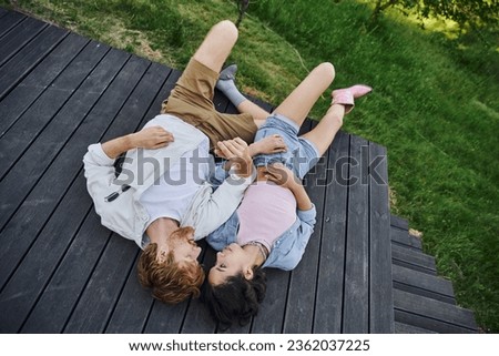 carefree multiethnic couple in stylish casual attire lying down on wooden porch outdoors, top view Royalty-Free Stock Photo #2362037225