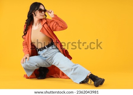 Stylish young Asian woman in fall clothes sitting on yellow background