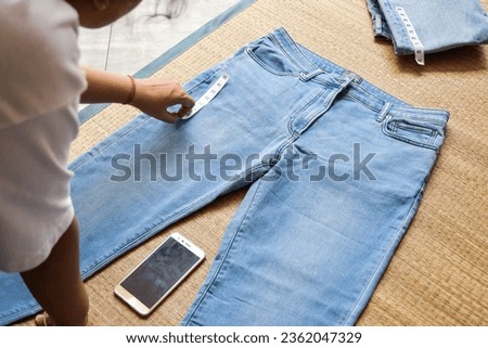 Woman hand attaching size labels on blue jeans on korai grass mat background. Preparation for livestream selling online or vlogging. 