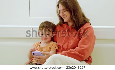 The child and his mother are waiting for their doctor's visit in the hospital lobby, scrolling through their phones together. Kid boy aged two years (two-year-old)
